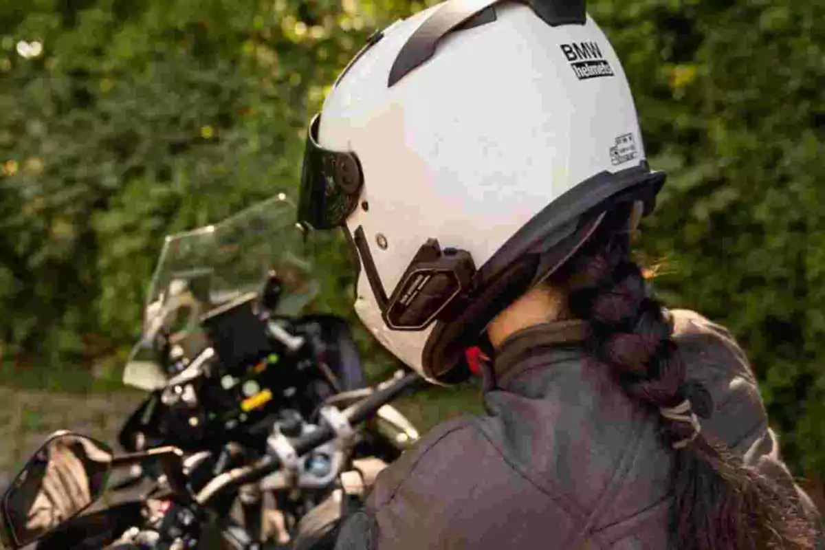 Persona con casco blanco y chaqueta oscura sentada en una motocicleta frente a un fondo de vegetación Persona con casco blanco y chaqueta oscura sentada en una motocicleta frente a un fondo de vegetación