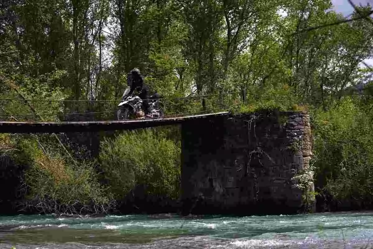 Person crossing a narrow bridge over a river on a motorcycle, surrounded by trees and vegetation Person crossing a narrow bridge over a river on a motorcycle, surrounded by trees and vegetation