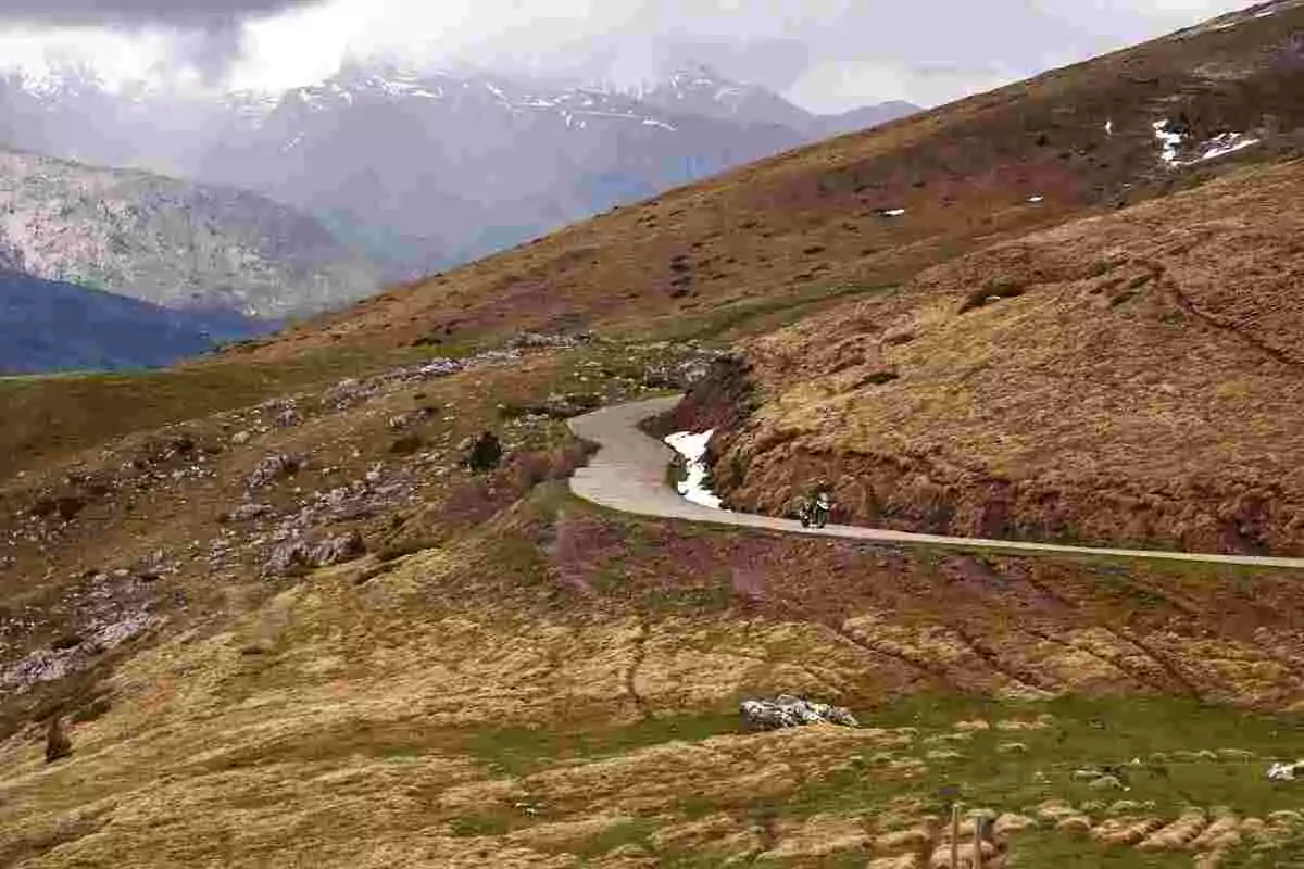 Winding mountain road between grass-covered hills and rocks with a motorcycle riding and snow-capped mountains in the background Winding mountain road between grass-covered hills and rocks with a motorcycle riding and snow-capped mountains in the background