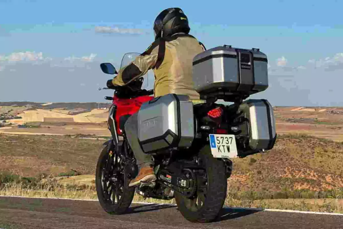 Persona con casco y chaqueta beige conduciendo una motocicleta roja equipada con maletas de viaje en una carretera con paisaje de campo al fondo Persona con casco y chaqueta beige conduciendo una motocicleta roja equipada con maletas de viaje en una carretera con paisaje de campo al fondo