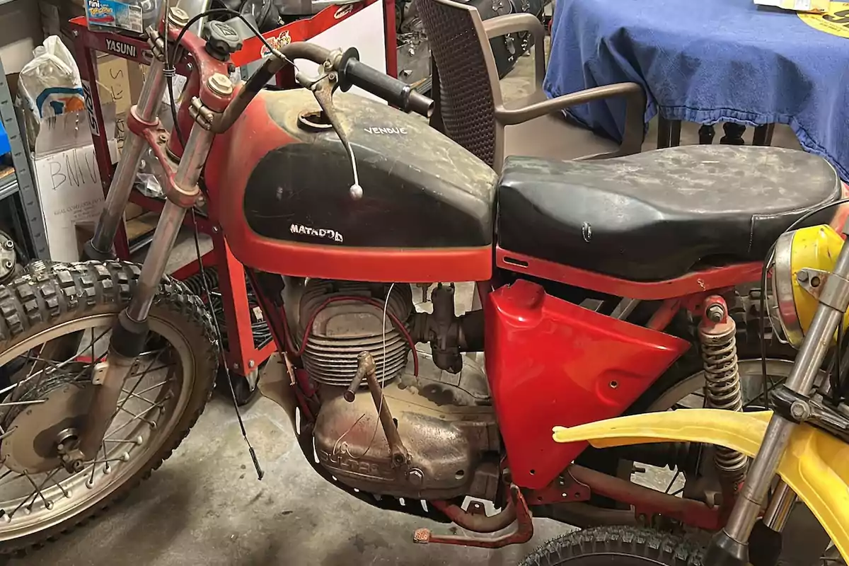 Motocicleta clásica Bultaco Matador en un taller con polvo sobre el asiento y el tanque, rodeada de herramientas y otros objetos Motocicleta clásica Bultaco Matador en un taller con polvo sobre el asiento y el tanque, rodeada de herramientas y otros objetos