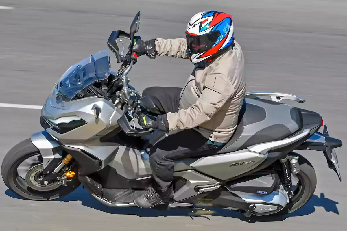 Persona conduciendo una motocicleta tipo scooter plateada en una carretera, usando casco y chaqueta protectora Persona conduciendo una motocicleta tipo scooter plateada en una carretera, usando casco y chaqueta protectora