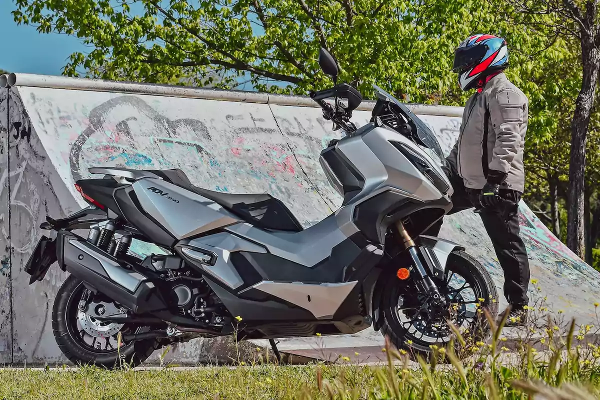 Motocicleta gris estacionada junto a una rampa de skate con grafitis y una persona con casco y chaqueta de protección de pie al lado Motocicleta gris estacionada junto a una rampa de skate con grafitis y una persona con casco y chaqueta de protección de pie al lado
