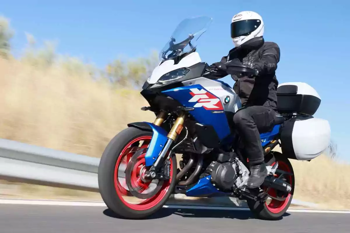Persona conduciendo una motocicleta deportiva BMW azul con maletas laterales blancas en una carretera bajo un cielo despejado