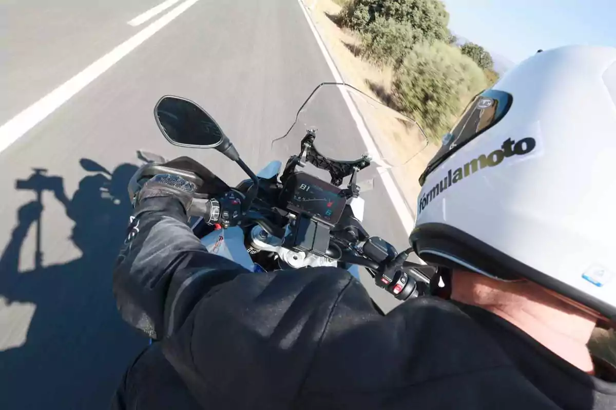 Persona conduciendo una motocicleta en carretera con casco blanco y chaqueta negra, vista desde arriba y ligeramente detrás Persona conduciendo una motocicleta en carretera con casco blanco y chaqueta negra, vista desde arriba y ligeramente detrás