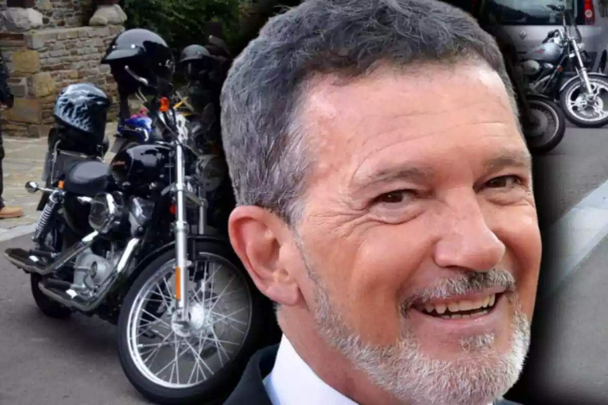 Un hombre de cabello canoso sonríe en primer plano, que es Antonio Banderas, mientras al fondo se ven varias motocicletas estacionadas en la calle.