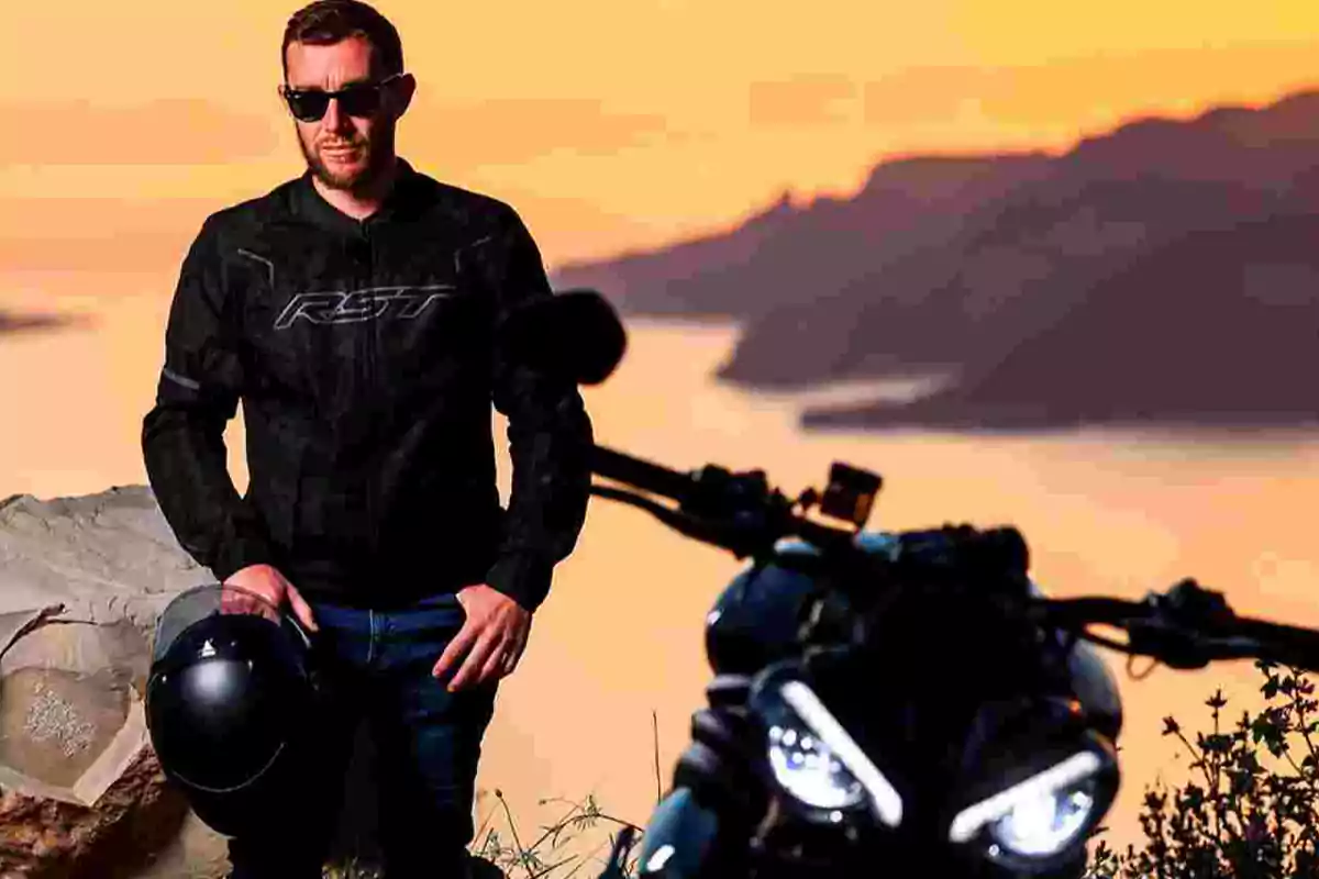 Hombre con chaqueta negra y gafas de sol sosteniendo un casco junto a una motocicleta al atardecer con paisaje de fondo