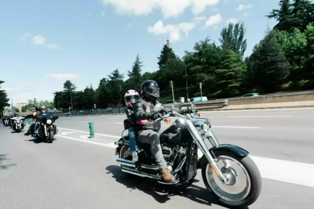 Dos personas con casco y chaqueta de cuero montan una motocicleta en una carretera junto a otros motociclistas en un día soleado Dos personas con casco y chaqueta de cuero montan una motocicleta en una carretera junto a otros motociclistas en un día soleado