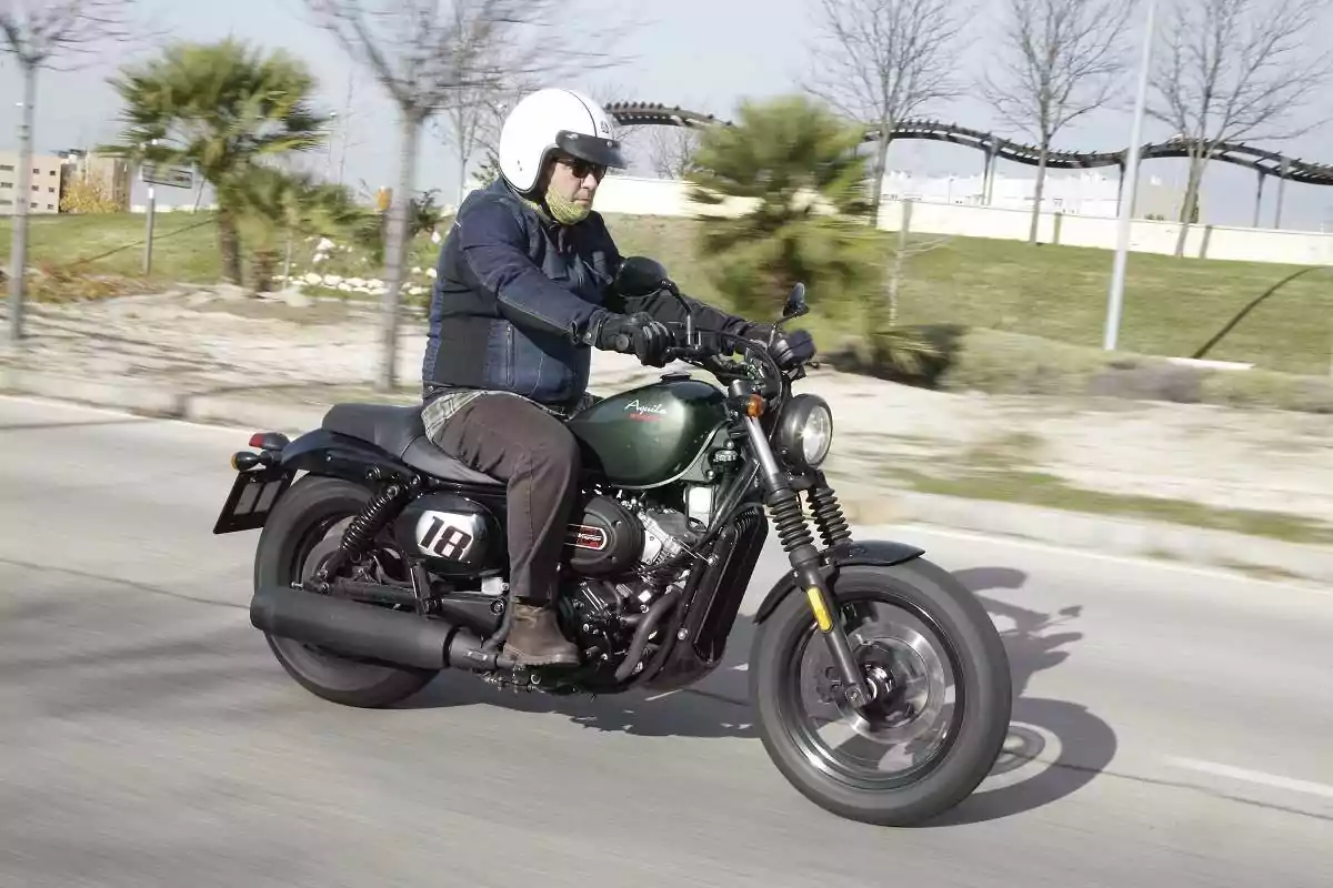 Hombre conduciendo una motocicleta verde en una calle con árboles y vegetación al fondo