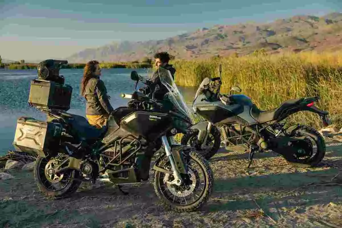 Dos personas conversan junto a dos motocicletas estacionadas cerca de un lago con montañas al fondo y vegetación alrededor Dos personas conversan junto a dos motocicletas estacionadas cerca de un lago con montañas al fondo y vegetación alrededor