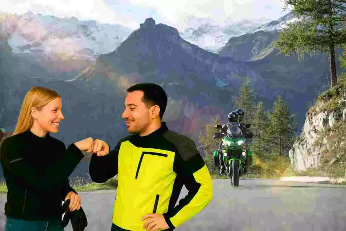 Dos personas con ropa de motociclista se saludan con el puño mientras una motocicleta se acerca por una carretera de montaña rodeada de árboles y montañas nevadas al fondo