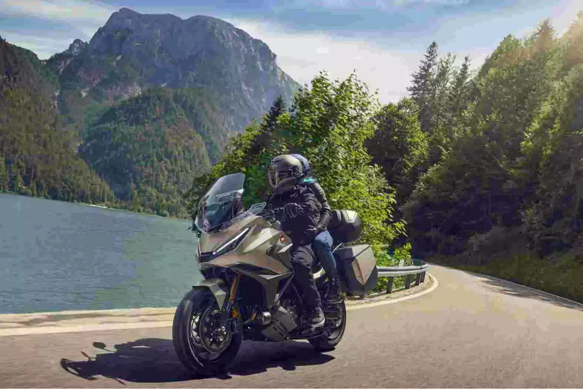 Dos personas viajan en motocicleta por una carretera junto a un lago rodeado de montañas y árboles Dos personas viajan en motocicleta por una carretera junto a un lago rodeado de montañas y árboles