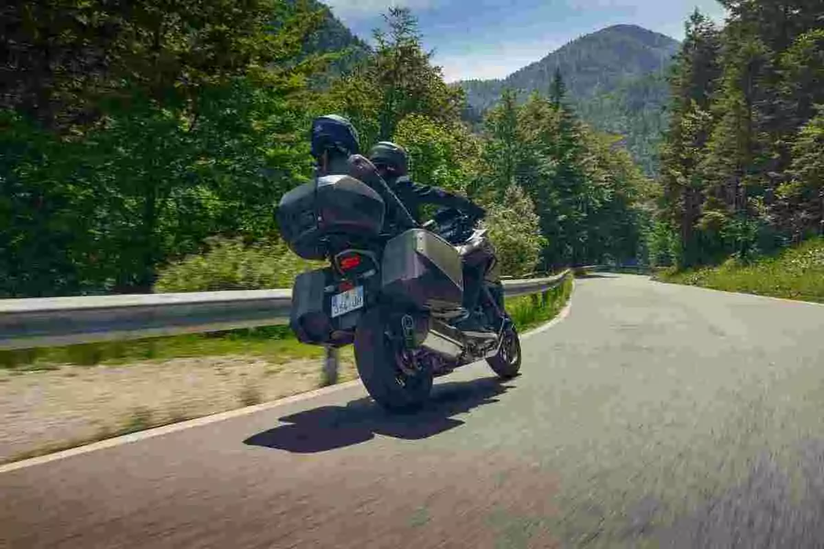 Dos personas en motocicleta recorren una carretera rodeada de árboles y montañas en un día soleado Dos personas en motocicleta recorren una carretera rodeada de árboles y montañas en un día soleado