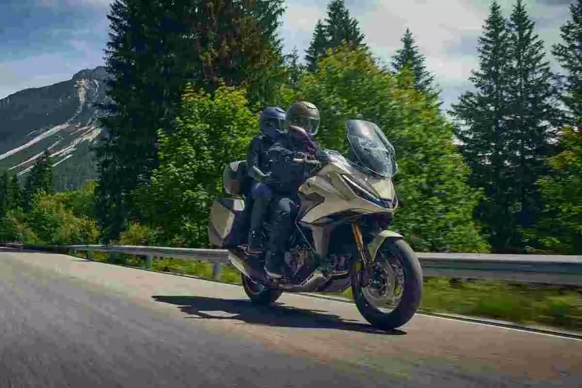 Dos personas con casco viajan en una motocicleta por una carretera rodeada de árboles y montañas. Dos personas con casco viajan en una motocicleta por una carretera rodeada de árboles y montañas.