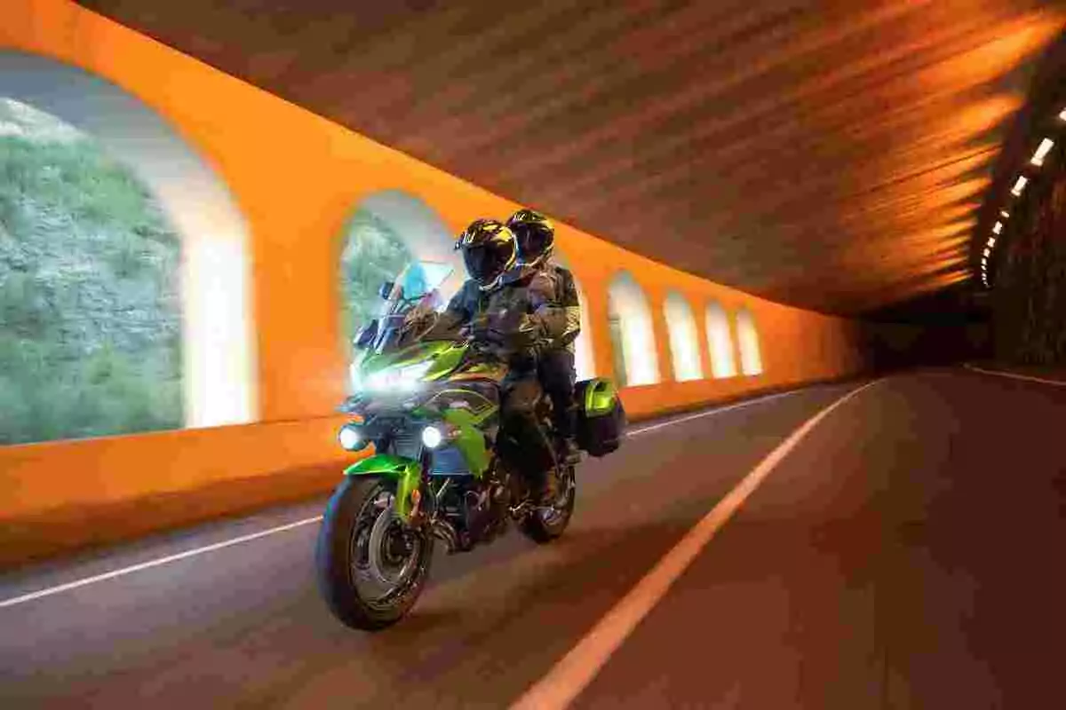 Dos personas con casco y equipo de protección viajan en una motocicleta verde dentro de un túnel iluminado con paredes naranjas y ventanas al exterior Dos personas con casco y equipo de protección viajan en una motocicleta verde dentro de un túnel iluminado con paredes naranjas y ventanas al exterior