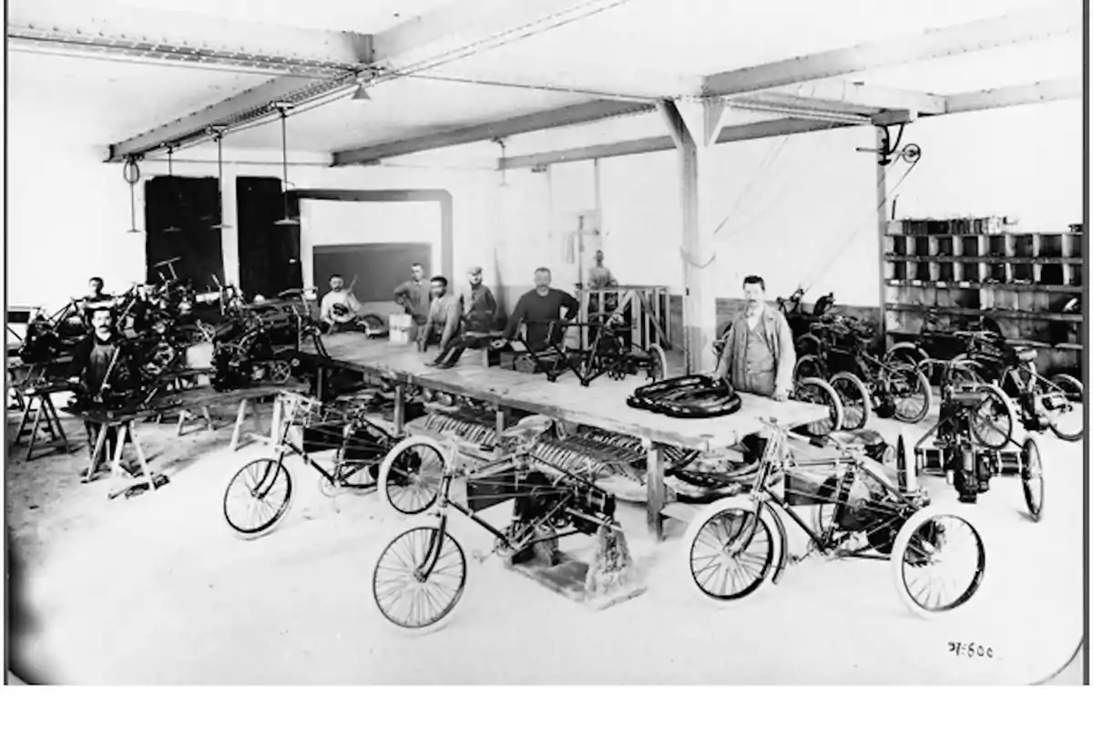 Varios hombres trabajan en un taller antiguo ensamblando triciclos y motocicletas rodeados de herramientas y piezas sobre mesas de madera Varios hombres trabajan en un taller antiguo ensamblando triciclos y motocicletas rodeados de herramientas y piezas sobre mesas de madera