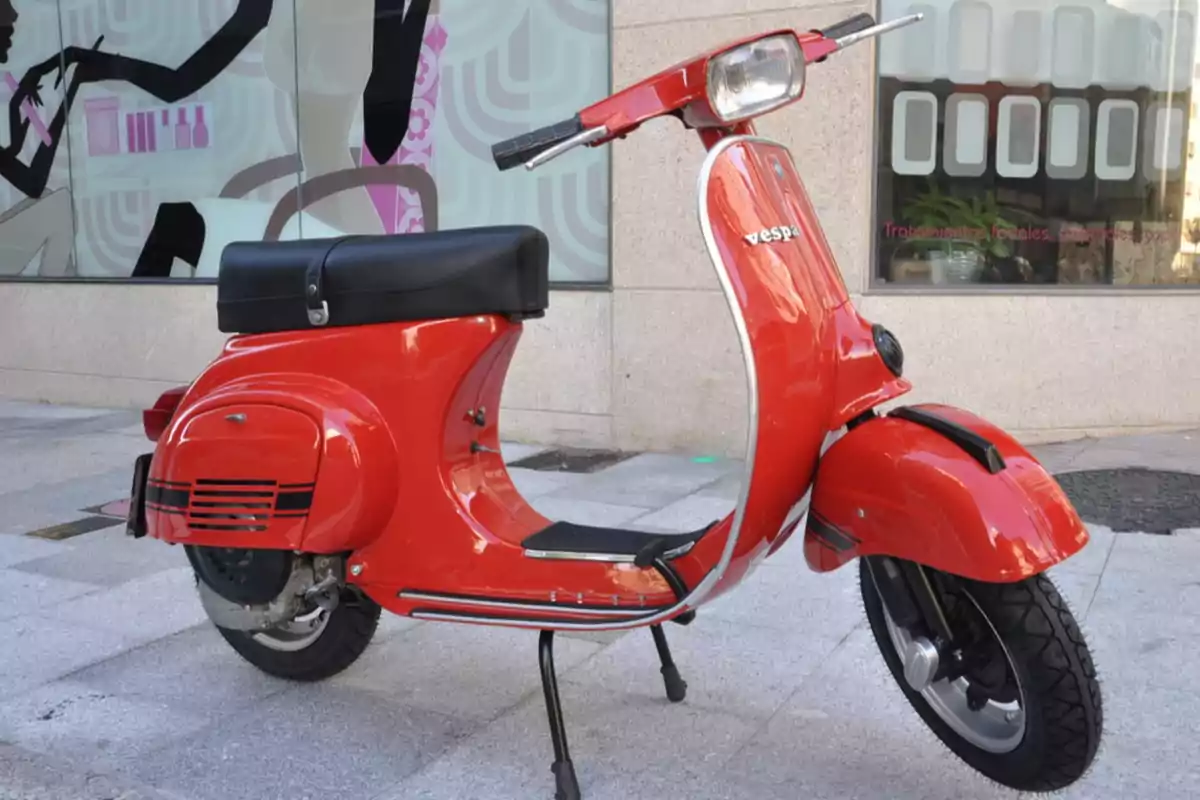 Motocicleta Vespa clásica de color rojo estacionada sobre una acera frente a un edificio con ventanas decoradas Motocicleta Vespa clásica de color rojo estacionada sobre una acera frente a un edificio con ventanas decoradas