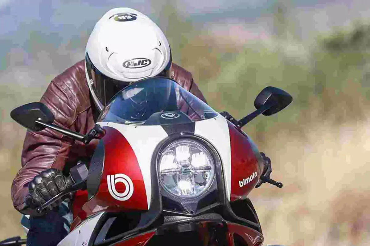 Persona con casco blanco y chaqueta marrón conduciendo una motocicleta deportiva roja y blanca de la marca Bimota en una carretera al aire libre Persona con casco blanco y chaqueta marrón conduciendo una motocicleta deportiva roja y blanca de la marca Bimota en una carretera al aire libre