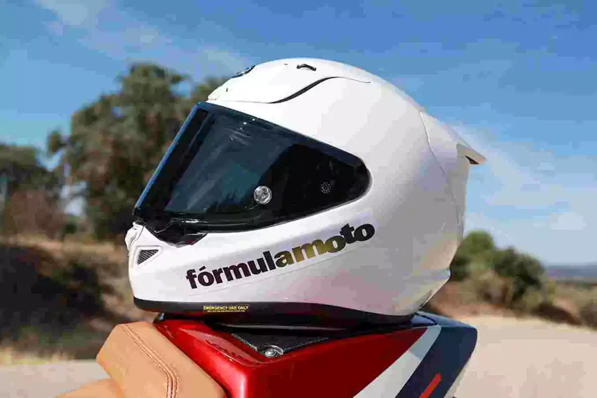 Casco de moto blanco con visera oscura y el texto fórmula moto en el lateral, apoyado sobre una motocicleta roja en un entorno al aire libre Casco de moto blanco con visera oscura y el texto fórmula moto en el lateral, apoyado sobre una motocicleta roja en un entorno al aire libre