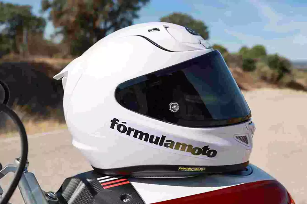 Casco de motocicleta blanco con visera oscura y el texto fórmulamoto en el lateral, apoyado sobre una moto en una carretera Casco de motocicleta blanco con visera oscura y el texto fórmulamoto en el lateral, apoyado sobre una moto en una carretera