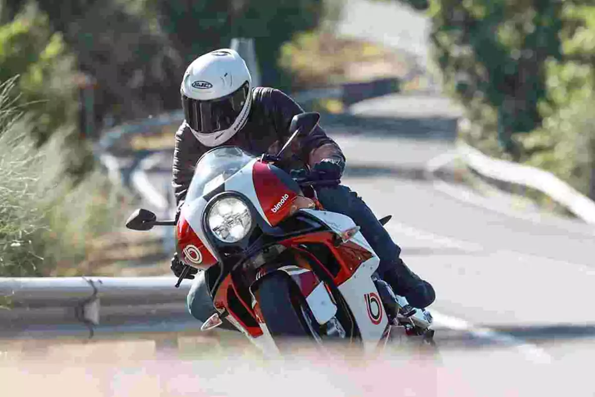 Persona con casco blanco conduciendo una motocicleta deportiva blanca y roja en una carretera rodeada de vegetación Persona con casco blanco conduciendo una motocicleta deportiva blanca y roja en una carretera rodeada de vegetación