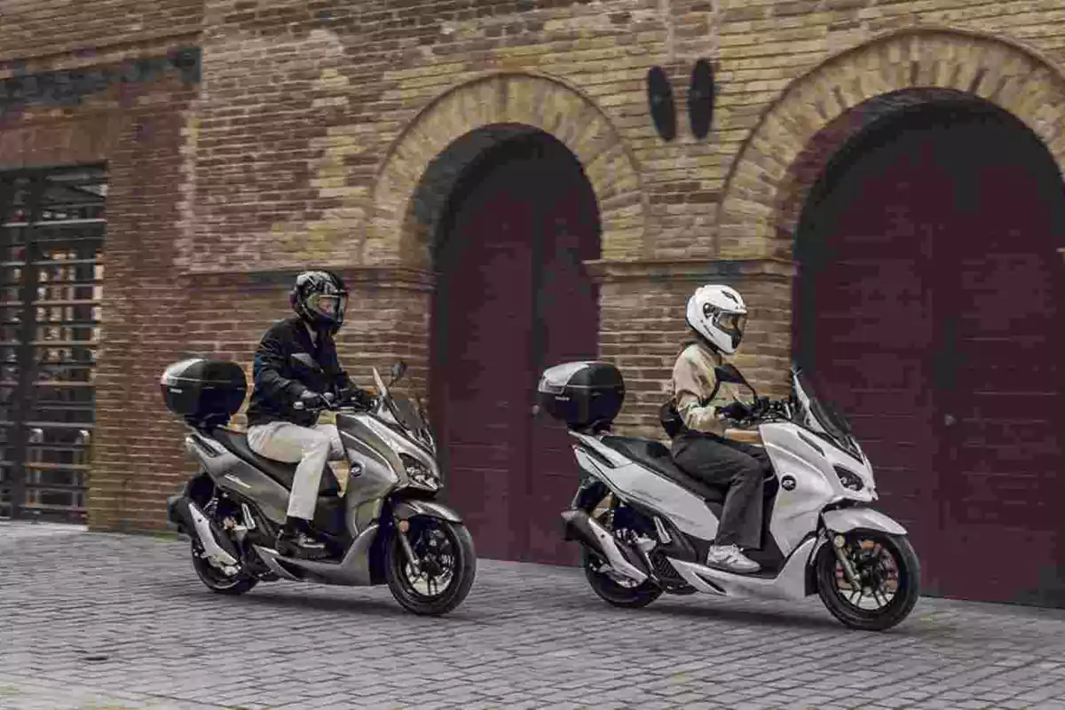 Dos personas con casco y chaqueta conduciendo scooters en una calle empedrada frente a un edificio de ladrillo con arcos grandes Dos personas con casco y chaqueta conduciendo scooters en una calle empedrada frente a un edificio de ladrillo con arcos grandes
