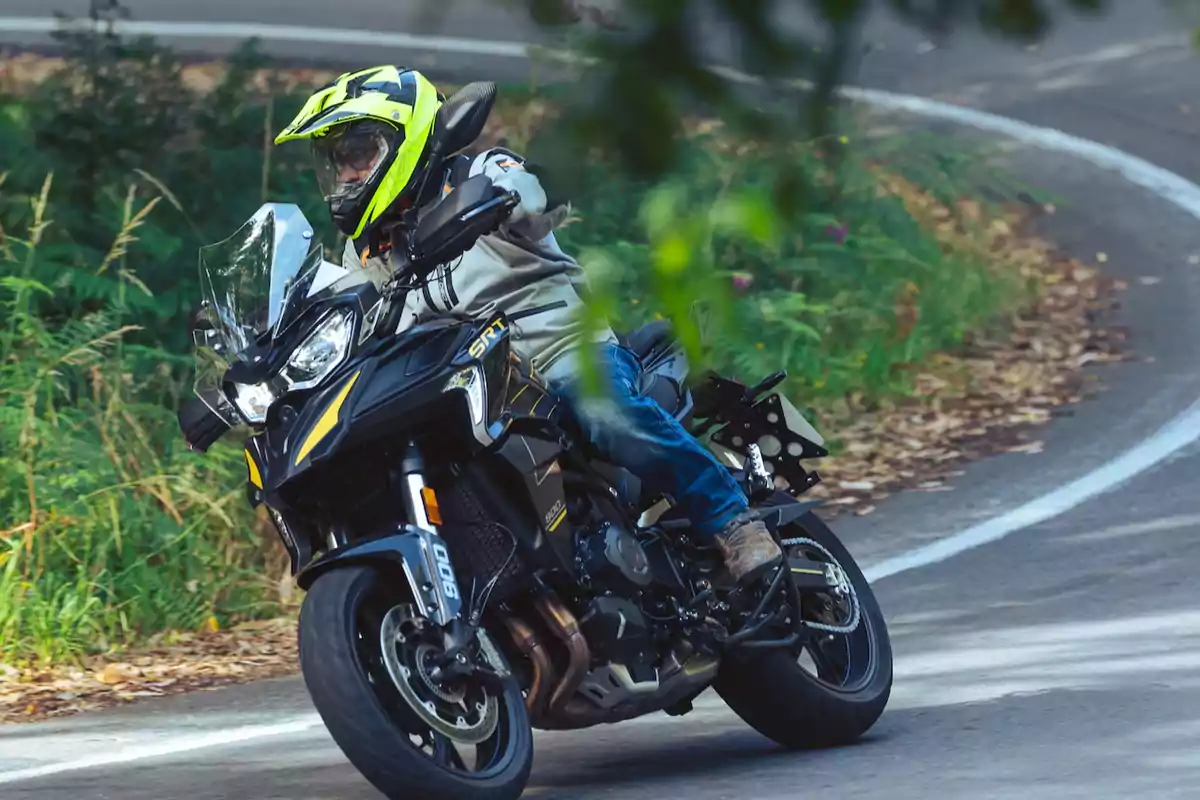 Persona con casco amarillo y chaqueta clara conduciendo una motocicleta negra en una curva de carretera rodeada de vegetación Persona con casco amarillo y chaqueta clara conduciendo una motocicleta negra en una curva de carretera rodeada de vegetación