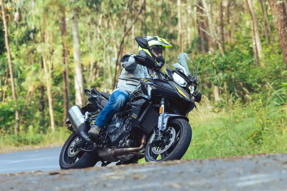 Persona con casco y chaqueta conduciendo una motocicleta deportiva negra en una carretera rodeada de árboles y vegetación Persona con casco y chaqueta conduciendo una motocicleta deportiva negra en una carretera rodeada de árboles y vegetación