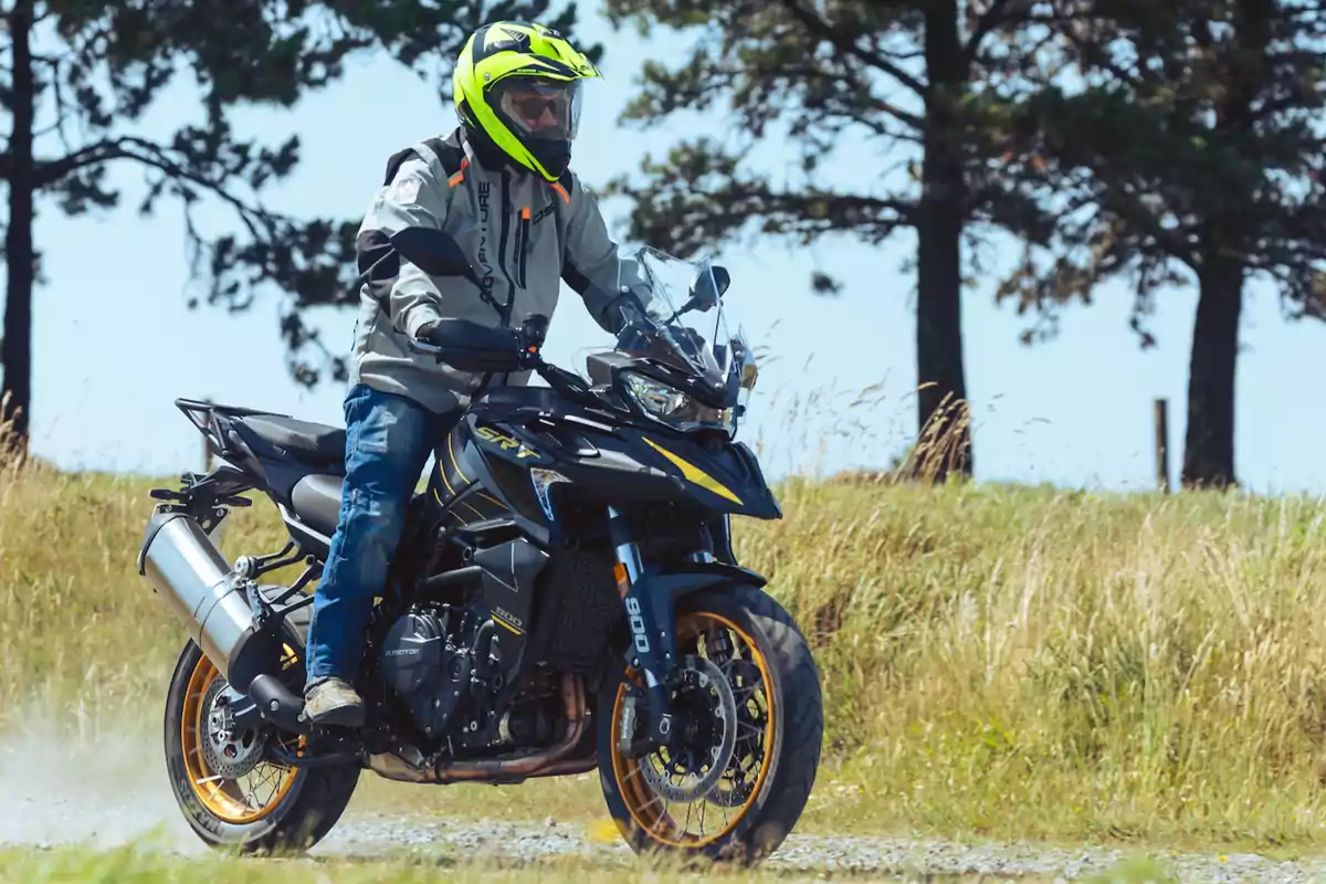Persona con casco amarillo y chaqueta gris conduciendo una motocicleta negra con detalles amarillos sobre un camino de tierra rodeado de pasto y árboles al fondo Persona con casco amarillo y chaqueta gris conduciendo una motocicleta negra con detalles amarillos sobre un camino de tierra rodeado de pasto y árboles al fondo