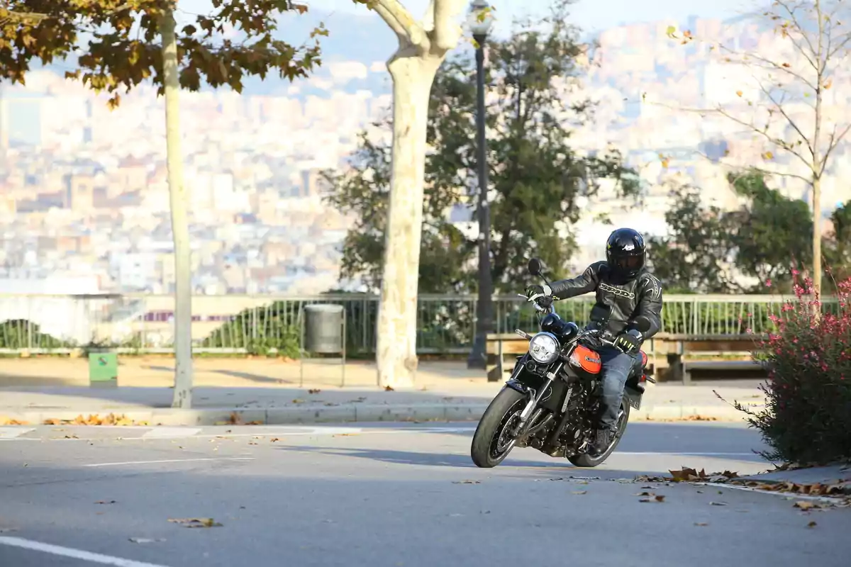 Persona con casco y chaqueta de motociclista conduciendo una moto en una calle con árboles y vegetación al fondo Persona con casco y chaqueta de motociclista conduciendo una moto en una calle con árboles y vegetación al fondo