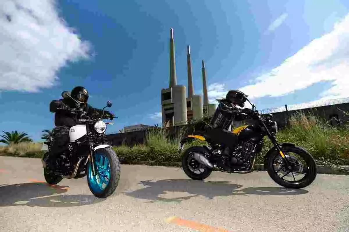 Dos personas con casco y ropa de protección montan motocicletas en una carretera con una central eléctrica de tres chimeneas al fondo bajo un cielo azul Dos personas con casco y ropa de protección montan motocicletas en una carretera con una central eléctrica de tres chimeneas al fondo bajo un cielo azul