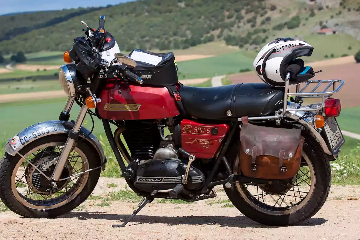 Motocicleta clásica roja estacionada en un camino rural con un casco blanco sobre el portaequipaje y una alforja de cuero en la parte trasera Motocicleta clásica roja estacionada en un camino rural con un casco blanco sobre el portaequipaje y una alforja de cuero en la parte trasera