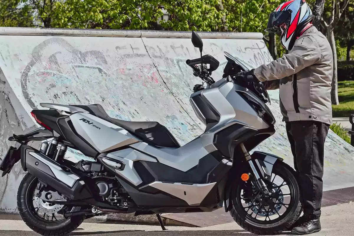 Persona con casco y chaqueta junto a una motocicleta plateada estacionada frente a una rampa de skate con grafitis Persona con casco y chaqueta junto a una motocicleta plateada estacionada frente a una rampa de skate con grafitis