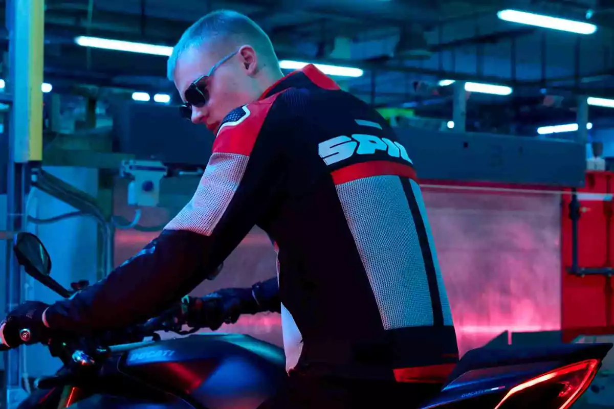 Hombre con gafas de sol y chaqueta de motociclista sentado en una moto Ducati en un entorno industrial iluminado con luces azules y rosas