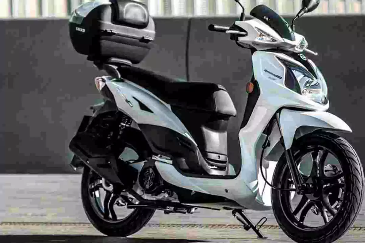 Motocicleta tipo scooter de color blanco estacionada sobre el pavimento con un baúl negro en la parte trasera