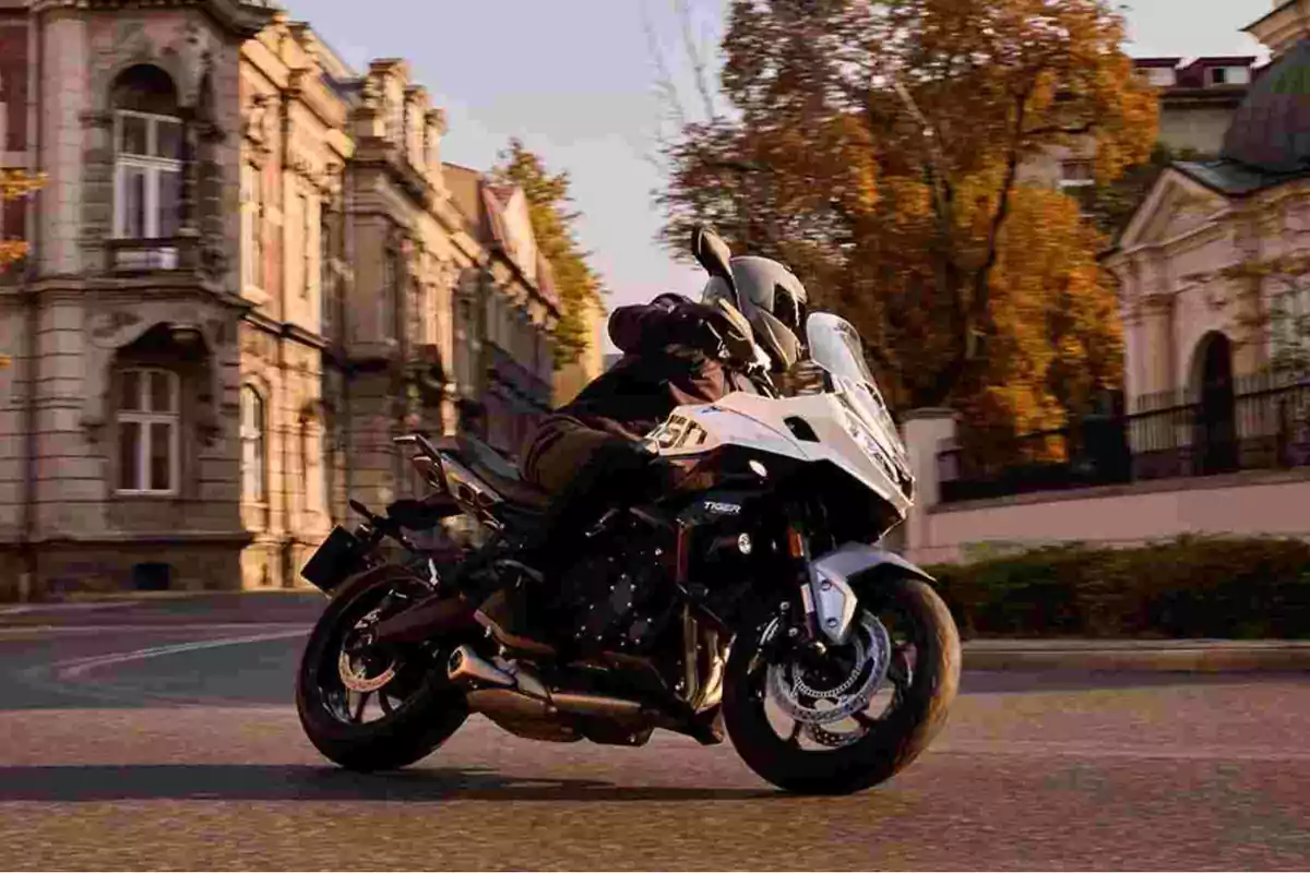Persona conduciendo una motocicleta deportiva blanca en una calle urbana con edificios antiguos y árboles al atardecer Persona conduciendo una motocicleta deportiva blanca en una calle urbana con edificios antiguos y árboles al atardecer