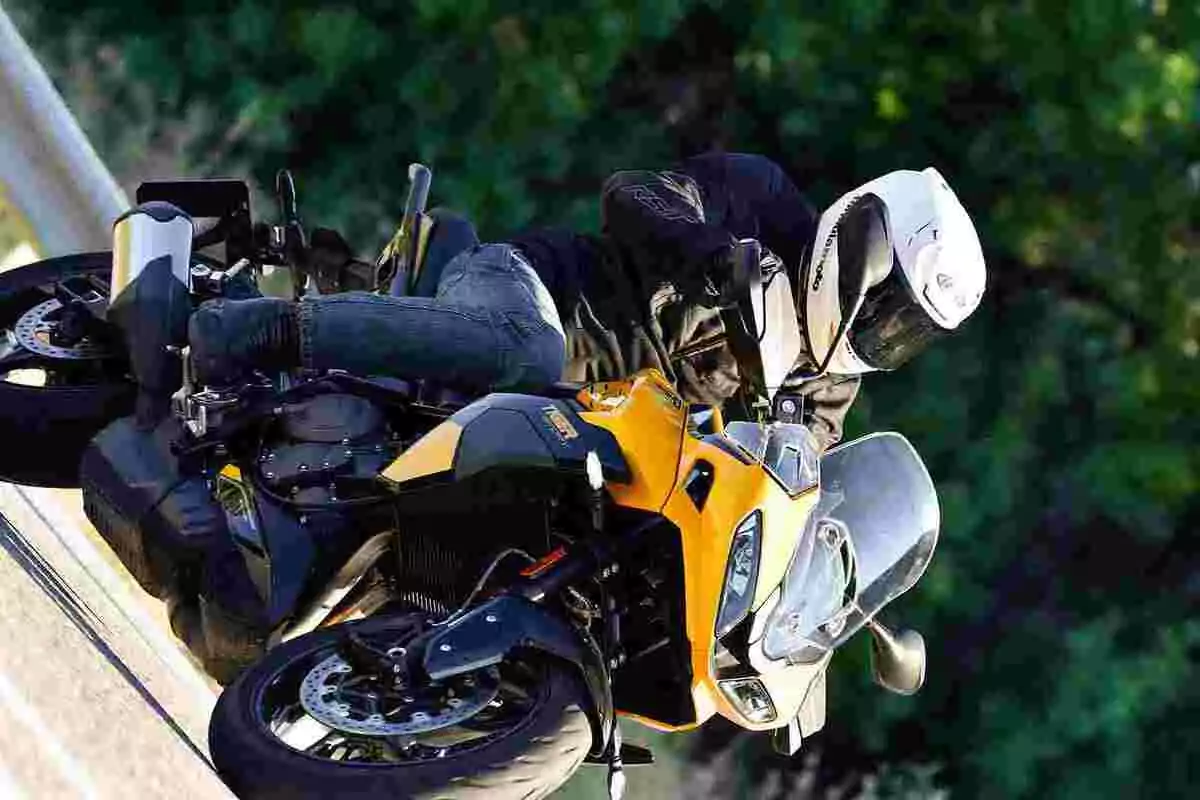 Persona con casco blanco y chaqueta negra conduciendo una motocicleta deportiva amarilla inclinada en una curva con fondo de vegetación verde Persona con casco blanco y chaqueta negra conduciendo una motocicleta deportiva amarilla inclinada en una curva con fondo de vegetación verde