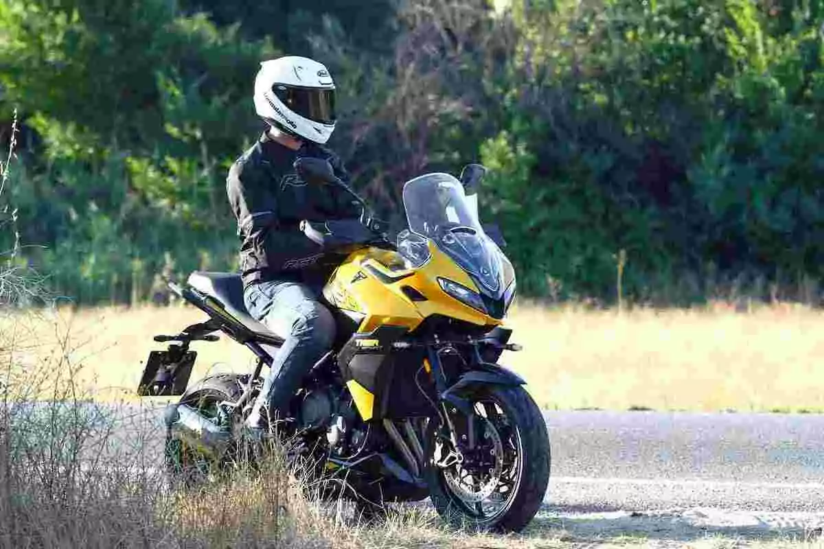 Persona con casco blanco y chaqueta negra sentada en una motocicleta deportiva amarilla junto a una carretera rodeada de vegetación Persona con casco blanco y chaqueta negra sentada en una motocicleta deportiva amarilla junto a una carretera rodeada de vegetación