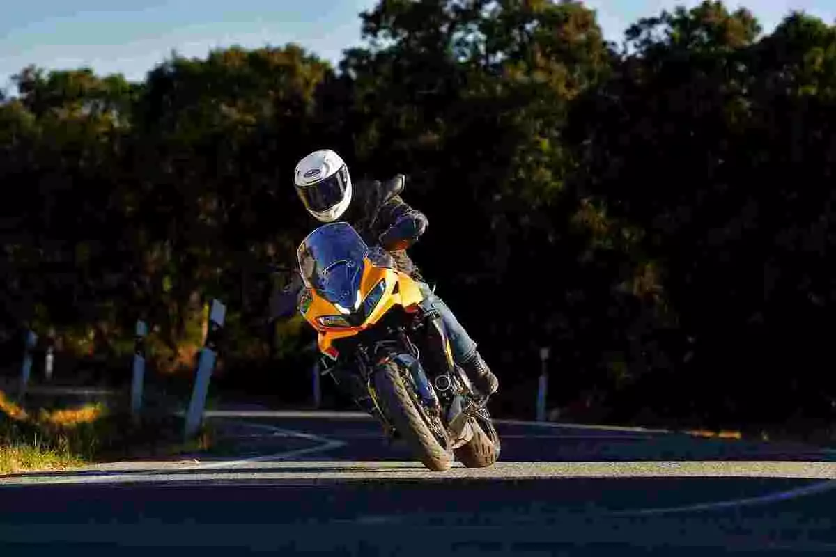 Persona con casco blanco conduciendo una motocicleta deportiva naranja en una carretera curva rodeada de árboles Persona con casco blanco conduciendo una motocicleta deportiva naranja en una carretera curva rodeada de árboles