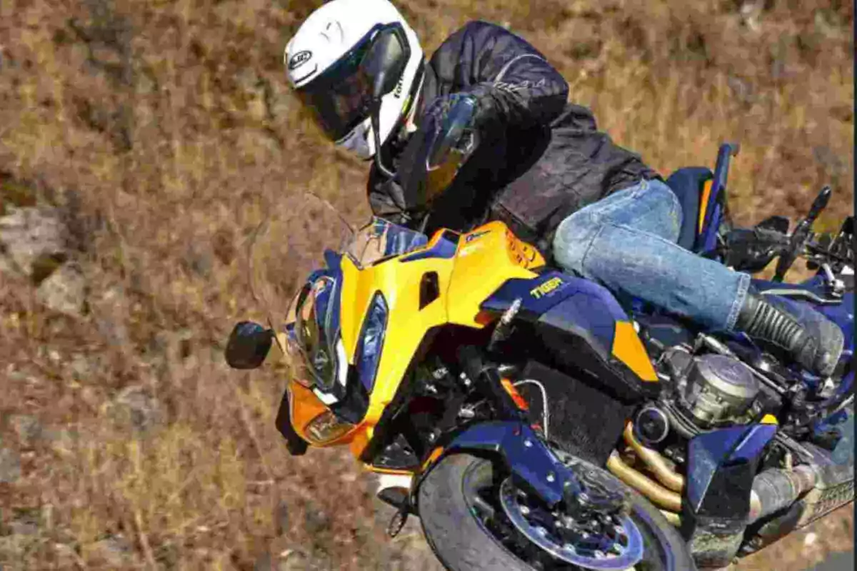 Persona con casco blanco y chaqueta negra conduciendo una motocicleta deportiva amarilla y azul en una carretera rodeada de vegetación seca