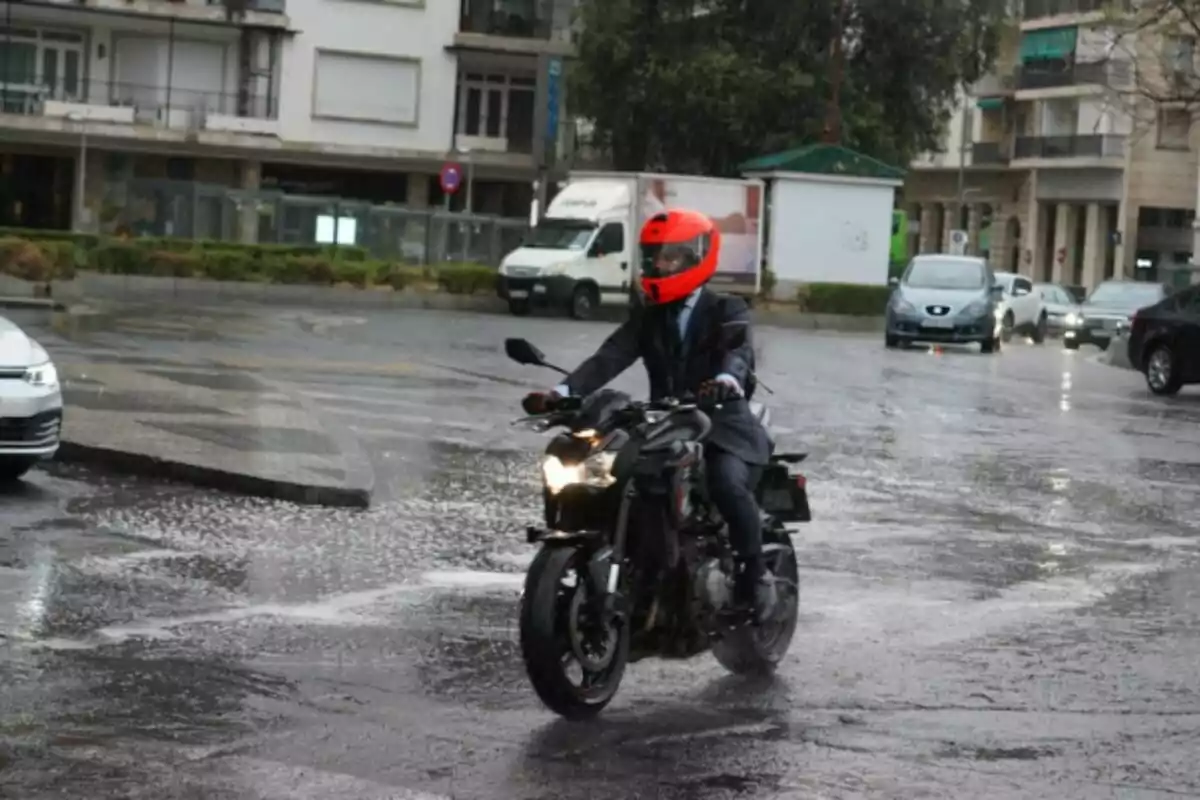 Persona con casco rojo conduciendo una motocicleta en una calle mojada por la lluvia rodeada de autos y edificios. Persona con casco rojo conduciendo una motocicleta en una calle mojada por la lluvia rodeada de autos y edificios.