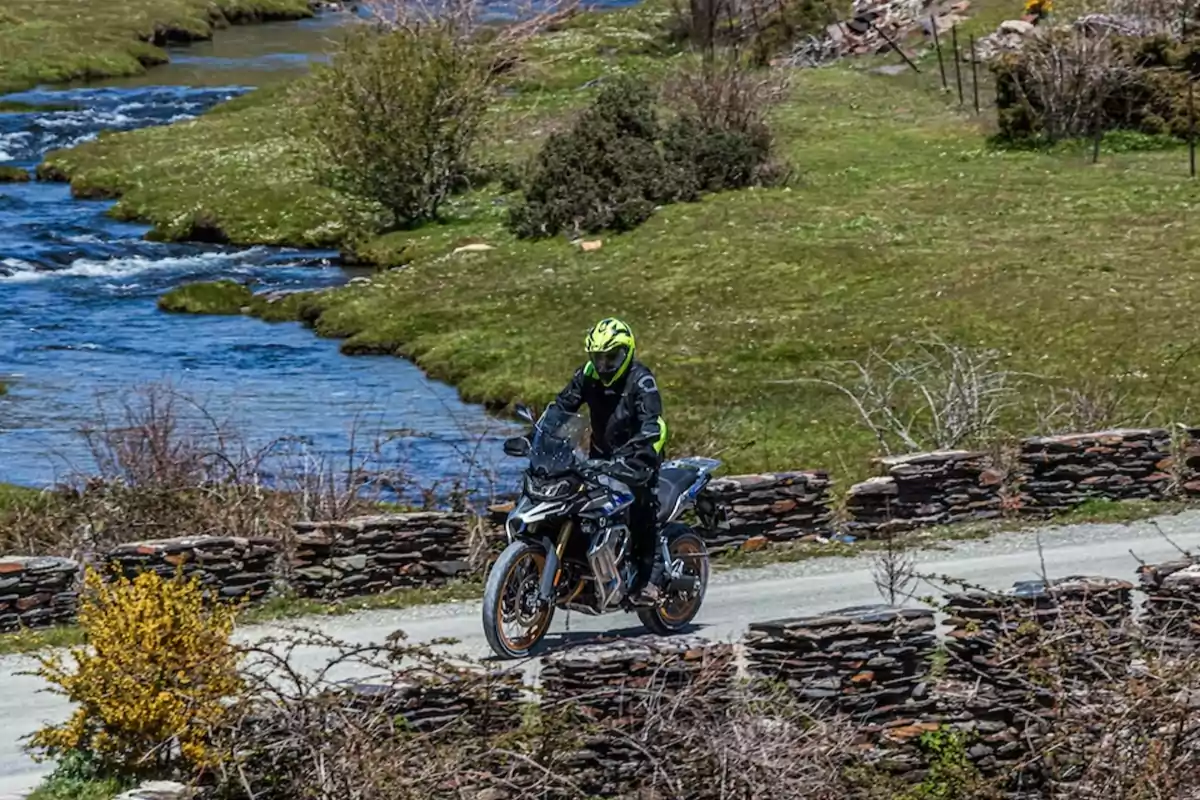 Persona conduciendo una motocicleta por un camino rural junto a un río rodeado de vegetación y muros de piedra Persona conduciendo una motocicleta por un camino rural junto a un río rodeado de vegetación y muros de piedra