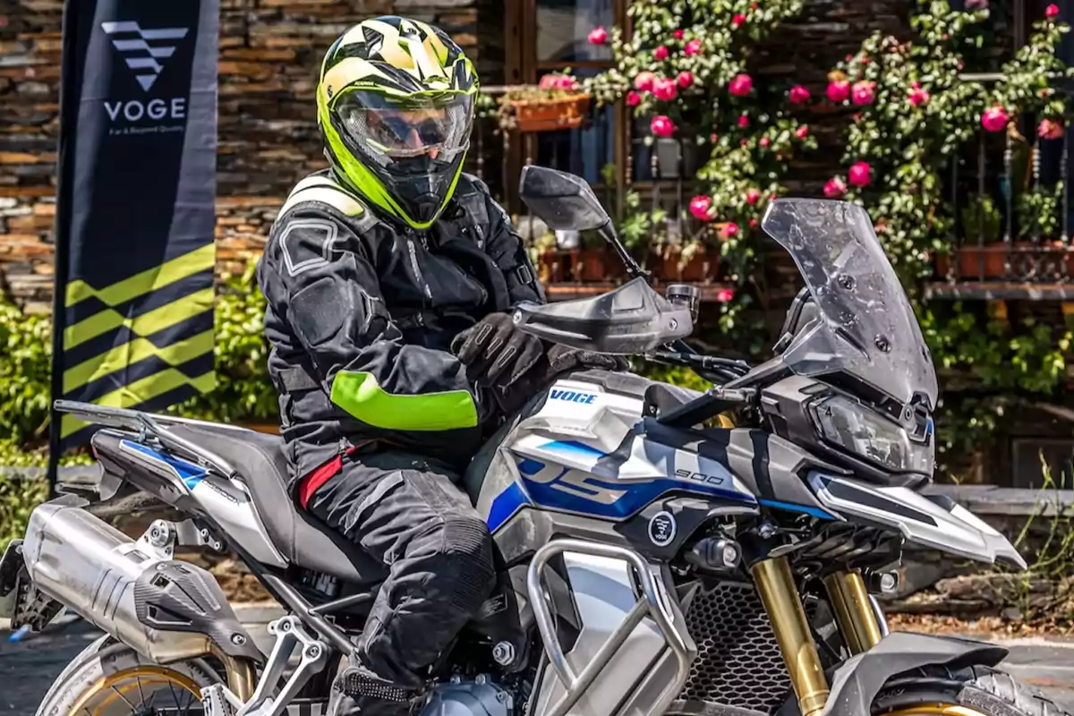 Persona con casco y equipo de protección negro y verde montada en una motocicleta Voge 900 DS estacionada al aire libre con flores y una bandera de la marca en el fondo Persona con casco y equipo de protección negro y verde montada en una motocicleta Voge 900 DS estacionada al aire libre con flores y una bandera de la marca en el fondo