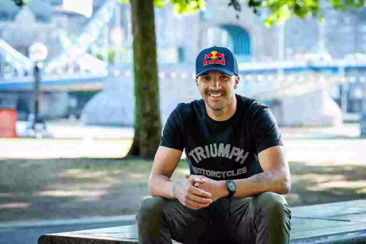 Hombre sonriente con gorra de Red Bull y camiseta de Triumph Motorcycles sentado en un banco al aire libre con un puente de fondo Hombre sonriente con gorra de Red Bull y camiseta de Triumph Motorcycles sentado en un banco al aire libre con un puente de fondo