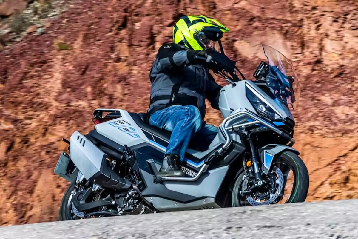 Persona conduciendo una motocicleta gris con casco amarillo en una carretera junto a una pared de roca