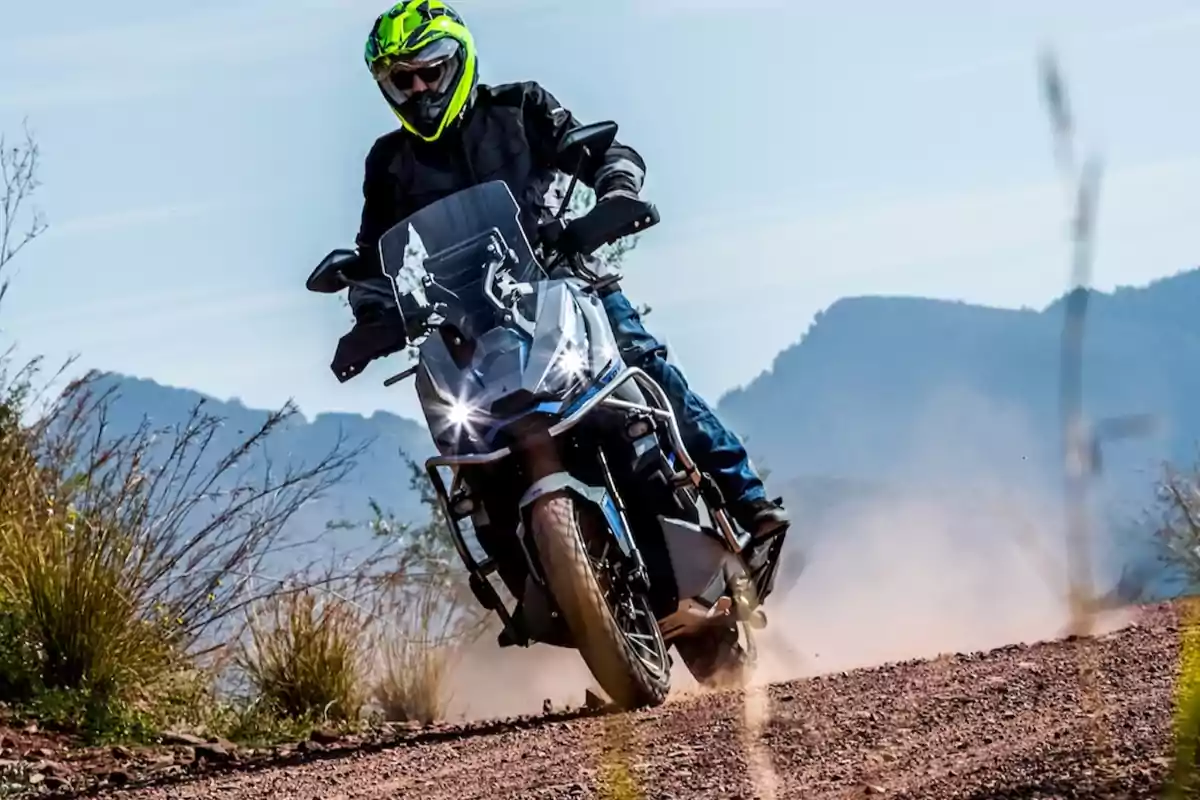 Persona con casco amarillo y chaqueta negra conduciendo una motocicleta en un camino de tierra con montañas al fondo Persona con casco amarillo y chaqueta negra conduciendo una motocicleta en un camino de tierra con montañas al fondo