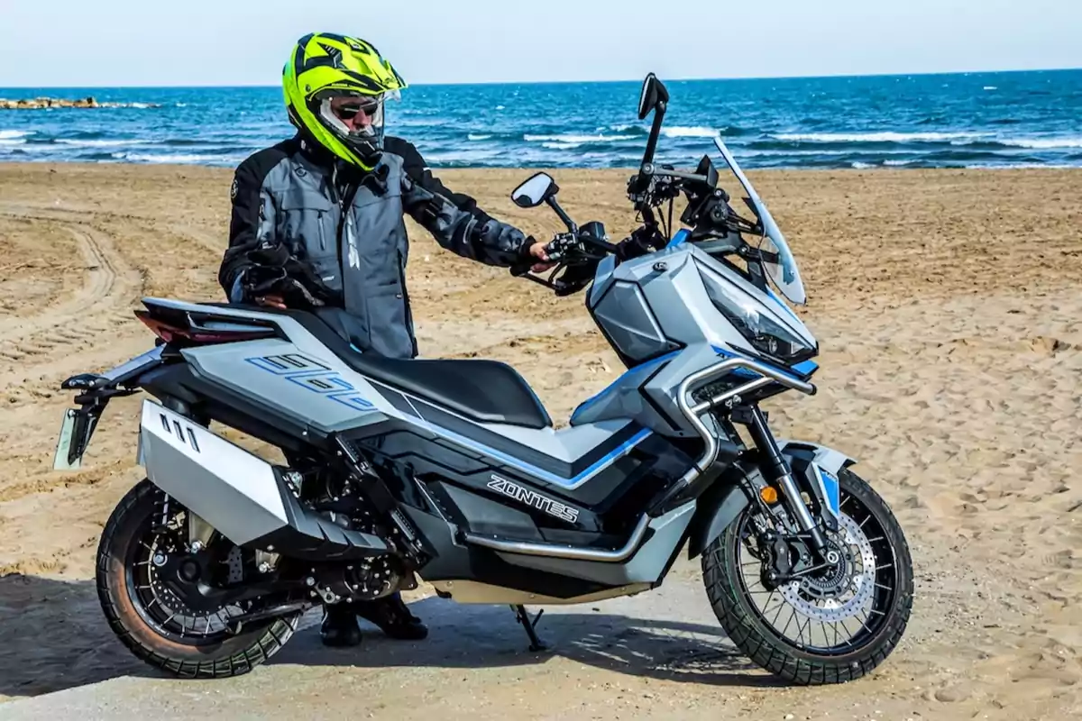 Persona con casco y chaqueta de motociclista junto a una moto gris en la playa con el mar de fondo Persona con casco y chaqueta de motociclista junto a una moto gris en la playa con el mar de fondo