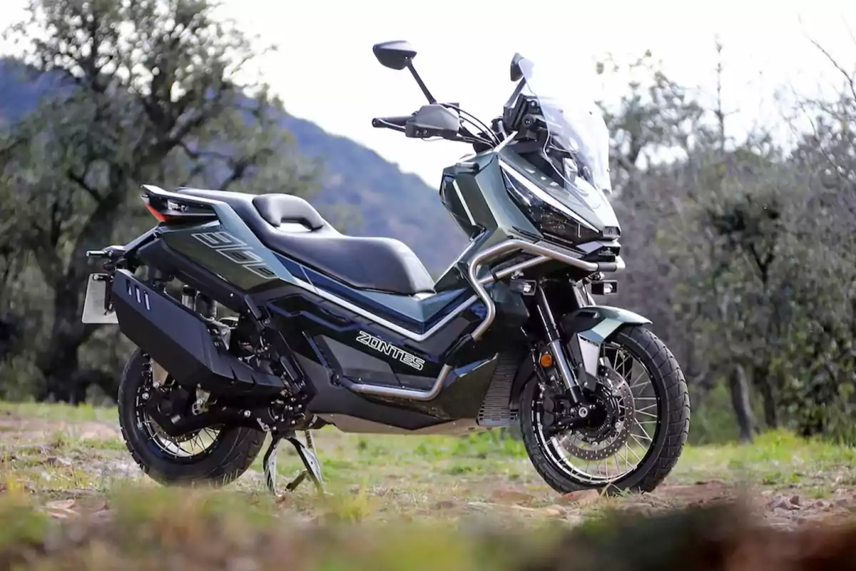 Motocicleta Zontes 310T estacionada al aire libre sobre un terreno de pasto y tierra con árboles y montañas al fondo Motocicleta Zontes 310T estacionada al aire libre sobre un terreno de pasto y tierra con árboles y montañas al fondo
