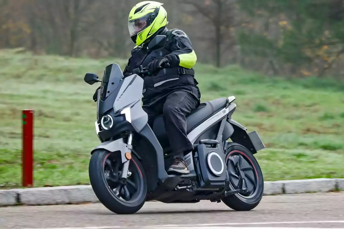 Person riding a gray electric motorcycle with a helmet and protective jacket on a road next to a green area