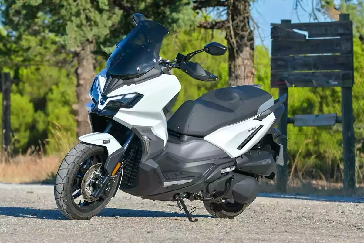 Motocicleta tipo scooter de color blanco estacionada en un camino de tierra con árboles y un letrero de madera al fondo Motocicleta tipo scooter de color blanco estacionada en un camino de tierra con árboles y un letrero de madera al fondo